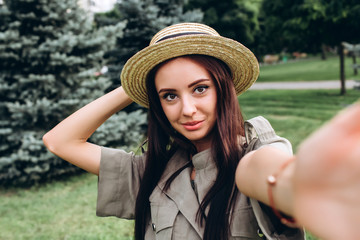 Young brunette hipster woman taking a selfie, showing tongue, funny face posing outdoors. Closeup portrait of a girl in a stylish summer hat in a park.