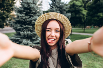 Young brunette hipster woman taking a selfie, showing tongue, funny face posing outdoors. Closeup portrait of a girl in a stylish summer hat in a park.