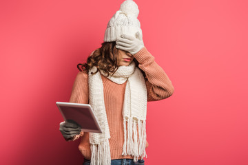 upset girl in warm hat and scarf holding digital tablet and covering face with hand on pink background