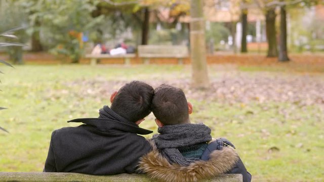 two young gays sitting on a bench at park approaching heads