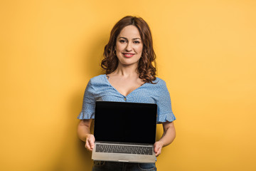 smiling woman holding laptop with blank screen on yellow background