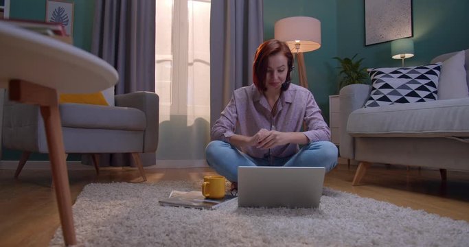 Young attractive Caucasian smiled woman with red hair speking with a mic via videochat on the laptop computer. At home.