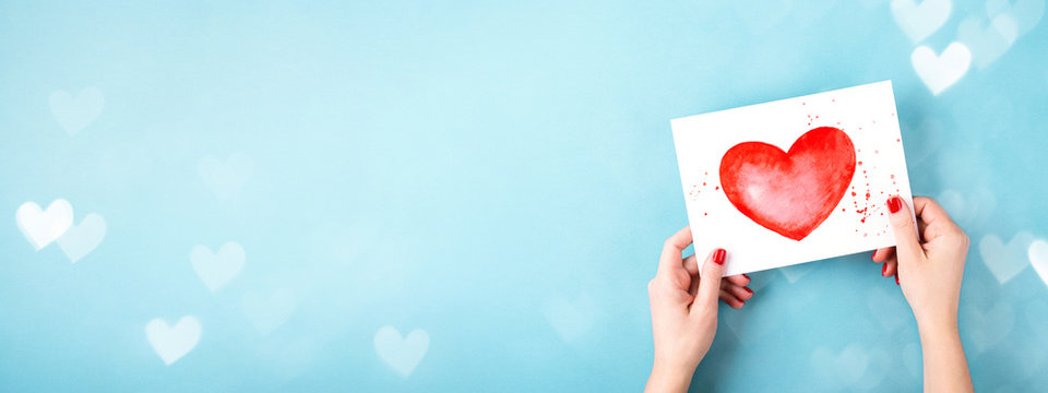 Female hands holding card with a painted red heart on blue background with bokeh. Minimal styled banner for Valentine's day.