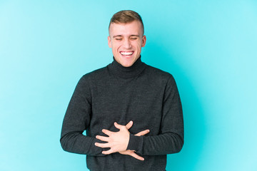 Young caucasian man on a blue background laughs happily and has fun keeping hands on stomach.