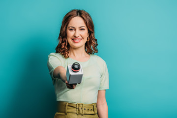 positive journalist smiling at camera while holding microphone on blue background - obrazy, fototapety, plakaty