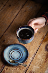 Closeup black americano coffee in blue cup in male hand, saucer with vintage tea spoon on wooden brown vintage background. Concept breakfast in coffee house