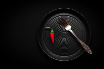 Closeup round ceramic plate with pattern of circles, vintage fork, red fresh chili pepper on black background. Concept modern shooting of advert menu from chef for a restaurant, tableware catalog