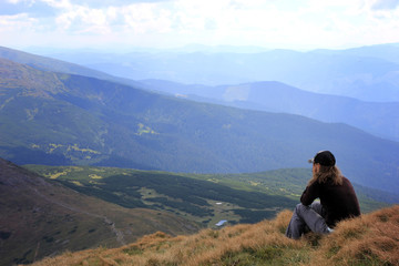 Girl in a cap sitting on a high mountain, looking at the mountains, road and clouds.