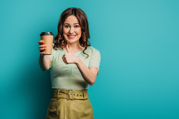 cheerful girl showing thumb up while holding coffee to go on blue background