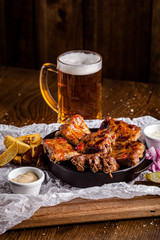 German cuisine concept. Baked pork ribs in marinade with a side dish of french fries, pickled onions, cucumbers and sauce. Serving in a cast-iron pan in restaurant on wooden table