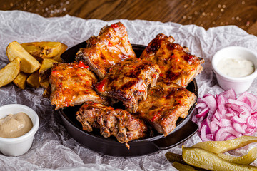 German cuisine concept. Baked pork ribs in marinade with a side dish of french fries, pickled onions, cucumbers and sauce. Serving in a cast-iron pan in restaurant on wooden table