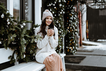 Winter portrait. A young woman with a stylish white sweater and a knitted hat poses against the background of Christmas decorations on a street in the city. The girl is drinking coffee in a Cup.