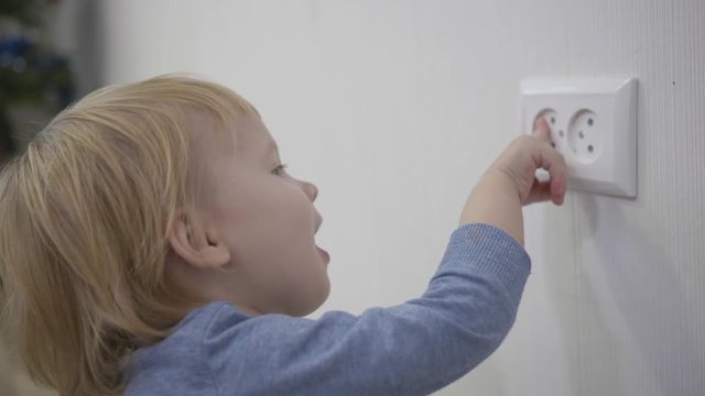 curious child is in danger, small boy tries to insert a finger into the electric socket close-up at home