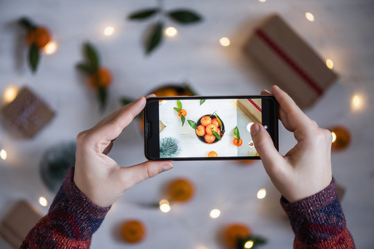 Woman blogger taking a picture on mobile phone her decoration of mandarin, Christmas tree, gifts, luminous garland on the white wooden boards background