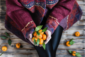 Woman in a huge winter sweater sits on the wooden rustic floor with small iron bucket of tangerines (mandarin).