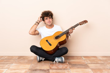 Young caucasian man with a guitar sitting on the floor having doubts and with confuse face...