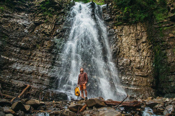 Traveler man with a yellow backpack standing on the background of a waterfall. Travel lifestyle concept.