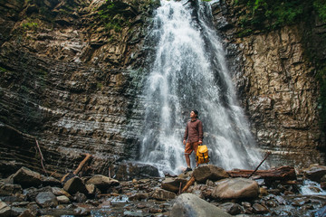 Traveler man with a yellow backpack standing on the background of a waterfall. Travel lifestyle concept.