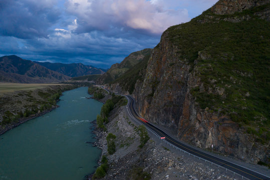 Aerial view of scenic Altai road between turquoise Katun River and rocky mountain wall at dusk. Highway P-256 Chuysky Trakt in summer evening. Nature landscape in Siberia, Russia