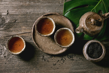 Hot black tea in traditional fireclay ceramic craft cups served with plate, teapot, exotic monstera leaf and bowl of dry tea leaves on old wooden table. Dark rustic atmosphere. Flat lay, space