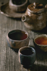 Hot black tea in different traditional wabi sabi style fireclay ceramic craft cups served with plate, teapot and bowl of dry tea leaves on old wooden table. Dark rustic atmosphere.