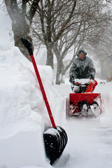 Man  pushing red snowblower in a February blizzard wearing winter gear and ear protection. Coat...