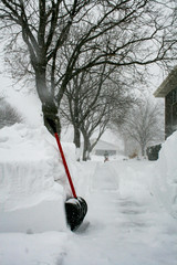 Red handled shovel prompt up against a 4 foot pile of snow along the sidewalk. Unidentified man in...