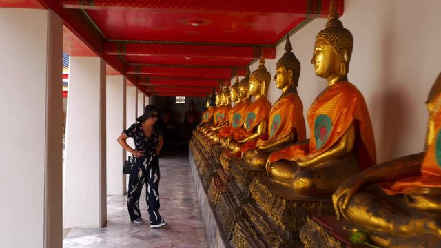 A woman walks past a row of Buddha statues in Wat Pho temple. She stops to egzamine some detail.