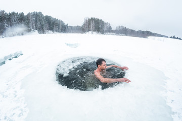 Young man swims in the ice hole made on the winter lake