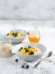Oatmeal porridge with mango, blueberries, and chopped almonds on white wooden table. Jar of honey on background. Selective focus. Superfoods, healthy breakfast image on a light background, copy space.