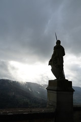 Statue of a Warrior with Speer looking over to the sunshine coming through clouds, hills surrounding the Hohenzollern castle in Bisingen Germany. The warrior or guardian is seen in a silhouette. 