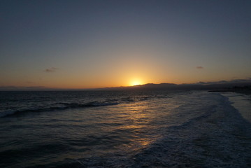 Fototapeta premium Sunsetting behind the mountains just off the beach of Santa Monica, California with a surfer and paraglider enjoying the views