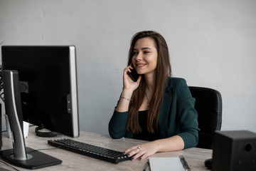 Portrait of young business woman talking phone against computer screen in white office. Female entrepreneur having call and working with computer sitting at table.Telephone work call.