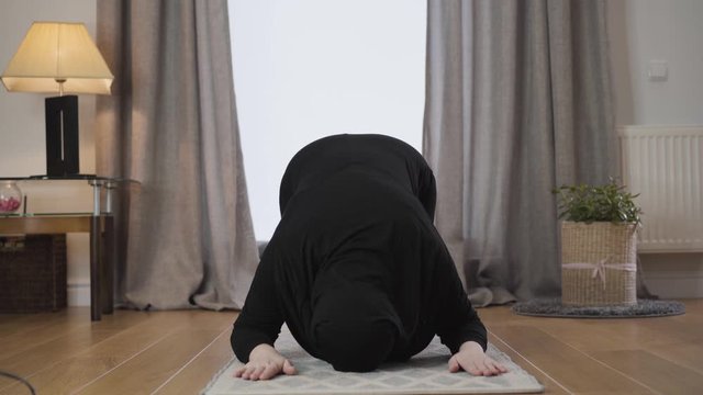 Portrait of young Muslim woman in black traditional dress and hijab praying at home. Beautiful lady looking up and bowing down. Eastern culture, religion, lifestyle.