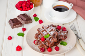 Chocolate waffles with caramel sauce, whipped cream and fresh raspberries with a cup of hot coffee on a white wooden background. Breakfast for Valentine's Day.
