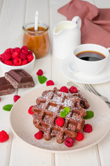 Chocolate waffles with caramel sauce, whipped cream and fresh raspberries with a cup of hot coffee on a white wooden background. Breakfast for Valentine's Day.