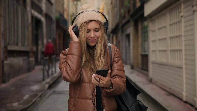Slow motion of young woman girl tourist in headphones and chocolate brown clothes dance on street and listening music from playlist on smartphone. Hipster girl with mobile phone in hands enjoying trip
