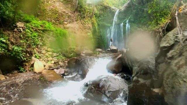 Slow motion, FPV, drone shot over people and rocks, towards the Sendang Gile waterfall, raindrops in frame, cloudy day, in Gunung Rinjani national park, Lombok, Indonesia