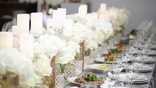 A festive table served by beautiful dishes in white colors surrounded by candles and peonies of flowers. Plates, cutlery and glasses for drinks.