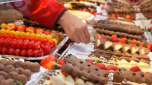 Various fruits on a wooden skewer stick in a caramelized glaze made of milk black white chocolate. Grapes, banana, strawberries in icing on the counter of the Christmas market