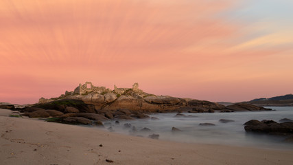 A ruin on a rock next to the Atalaia lighthouse in northern Spain in Galicia on the Atlantic. A long exposure at sunset with orange clouds and soft water.