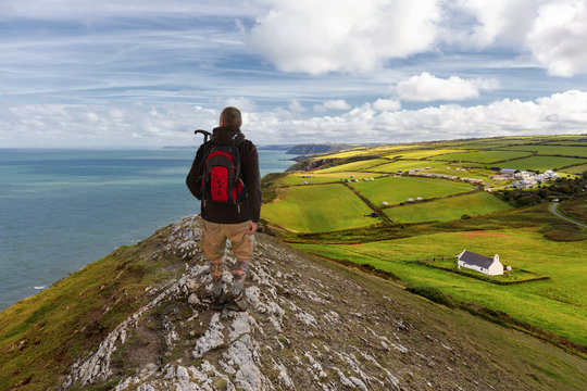 A hiker on the coastal path in Wales. The man looks at the sea. Below is the green land and a small white chapel. A cool and sunny day with a blue sky.