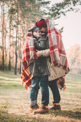 Couple huggings travelers covered with plaid in the forest. Concept of trekking, adventure and seasonal vacation.