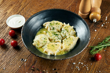 Boiled Ukrainian dumplings in butter with meat decorated with greens and sour cream in a black bowl on a wooden background. Tasty, traditional varenyky