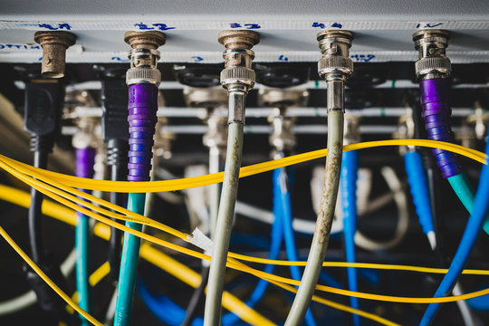 Blue BNC SDI signal cables plugged in the back of a patch panel viewed from top. A mess of cables visible behind the rack or panel. Focus on the first row of connectors.
