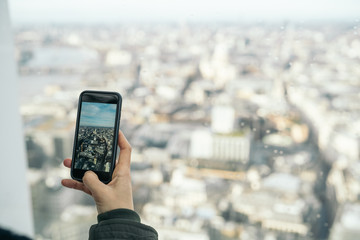 Woman taking a vertical photo with the smart phone of a panoramic view of the financial district of London City from the viewpoint of the Sky Garden.
