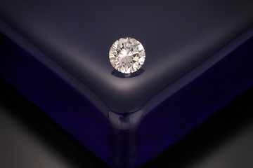 A solitaire, faceted, clear, round diamond sits on a reflective dark blue glass block background.