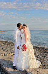 Fototapeta na wymiar Wedding by the sea. Beautiful couple of newlyweds. Bride with a red bouquet in a wedding dress at the water. The groom kisses the bride on the cheek. Concept marriage, just married.