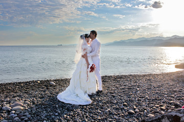 Fototapeta na wymiar Wedding by the sea. Lovely couple of newlyweds. Beautiful bride with a red bouquet. Mountains and sea background. Bride and groom kissing on stone beach during sunrise time. Just married in Batumi, Ge