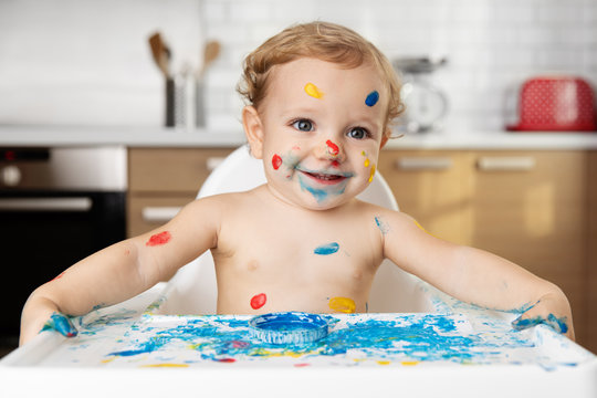 Cute smiling baby in high chair with finger paint on face and body